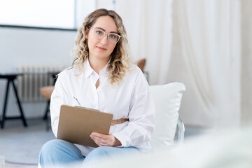 Close-up portrait of a young female therapist. Large glasses for vision, psychological help. Wavy blonde hair. There is a large light window in background.