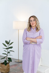 Portrait of a business woman in a purple shirt dress. She crossed her arms. Light and minimalistic interior. Looks into the camera.