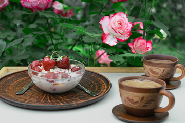 Delicious and healthy breakfast for two among the roses in the courtyard of the house. On a white table are two cups of coffee, strawberries with cottage cheese, cream and honey. Cloudy summer day