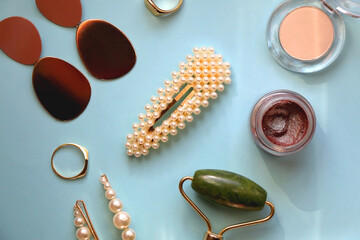 Various make-up products, jade face roller, pearl hair accessories, earrings and gold rings on bright blue background. Flat lay.