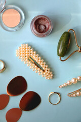 Various make-up products, jade face roller, pearl hair accessories, earrings and gold rings on bright blue background. Flat lay.