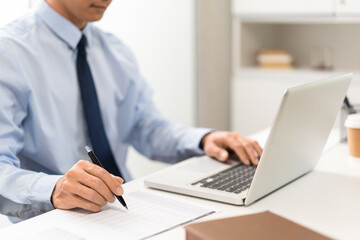 Obraz premium Working Man Conept The male officer sitting in the office, laying his left hand on the laptop, and writing something on the paper with his right hand