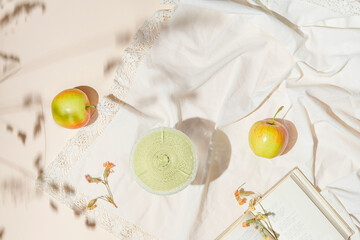 Creative composition made of glass with fruit juice, apples and book on pastel beige sunlit background with white cloth and shadows. Summer vacation and refreshment concept. Top view. Flat lay