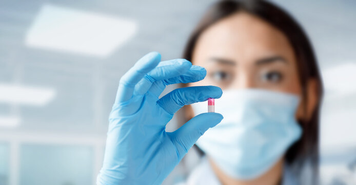 Female scientist wearing protective mask and gloves holds a capsule in pharmaceutical research lab.