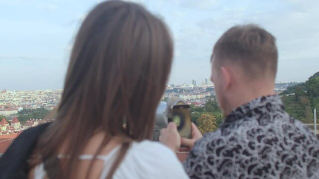 The backs of a guy and a girl on the panorama of the city. They stand near the viewing device. He is using the phone. Observation of the city from afar.