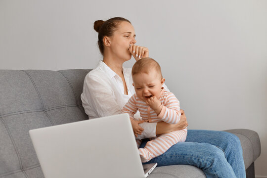 Image of sleepy female with bun hairstyle wearing white shirt and jeans sitting on sofa with her infant child and yawning, working long hours on laptop and looking after kid.