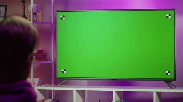 Man watching television in living room in evening. Young guy sitting on sofa against tv with chroma green screen, back view. Male person switching channels using remote controller.