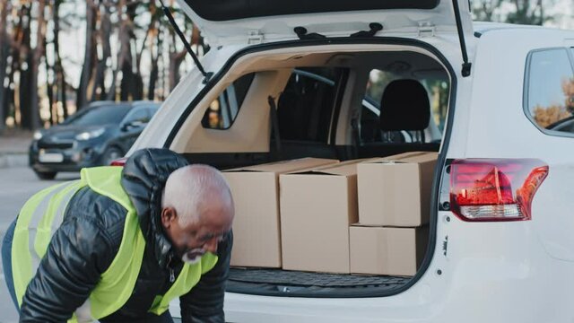 Close-up mature delivery service worker unloading cardboard boxes from trunk of car elderly african american courier loader delivering goods handyman smiling in work clothes accepts shipping outdoors