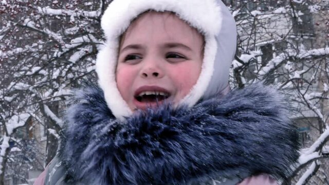 Portrait of little girl in frosty winter outdoors in snowfall. The child is warmly dressed in a gray fur collar, a hat with earflaps with white fur. Ruddy cheeks.