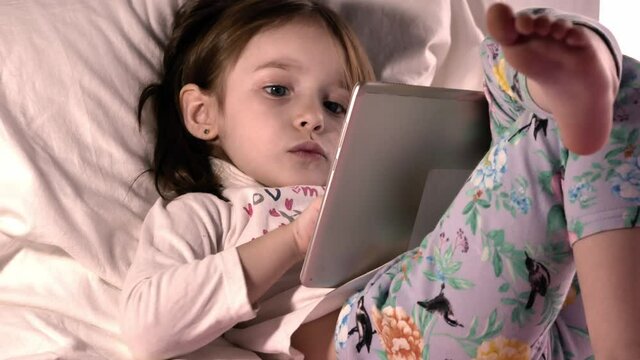 A little girl with straight disheveled hair sits on a French windowsill and watches an online lesson on a tablet or a cartoon or electronic game. Blue pants bare feet cozy in the morning at home.