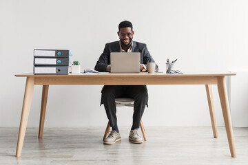 Cheerful African American businessman in suit working on laptop at office, full length