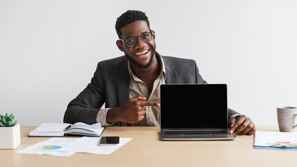 Happy young black businessman sitting at desk, pointing at laptop computer with blank screen at home office, mockup