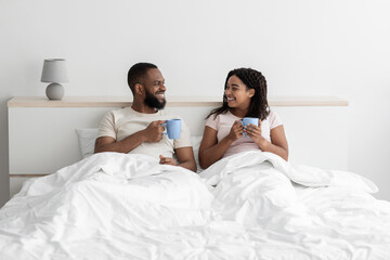 Smiling happy millennial african american man and wife sit on bed in white bedroom interior with cups of coffee