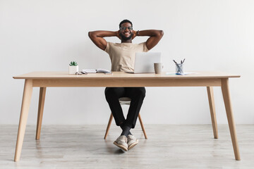 Obraz premium Happy millennial black man relaxing after work, putting feet on table at home office, full length