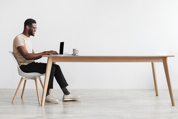 Side view of young black guy in casual wear using laptop, sitting at desk, working online from home...