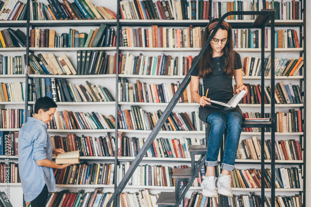 Wall mural Long haired teenage girl with glasses reads textbook preparing for tests while brunette woman takes book from shelf in spacious public library.