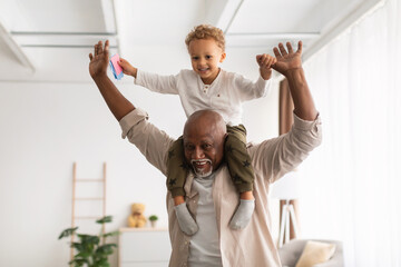 Cheerful Black Grandpa Carrying Little Grandson On Shoulders Playing Indoor - obrazy, fototapety, plakaty