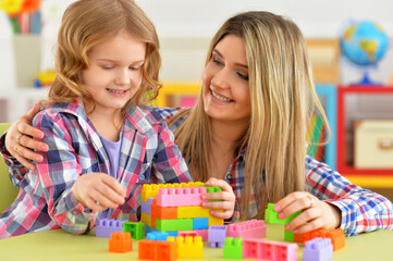 Naklejka premium Portrait of little girl and her mother playing colorful plastic blocks together in her room
