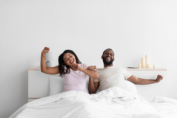 Cheerful young black boyfriend and girlfriend wake up after night sleep and stretch body on bed