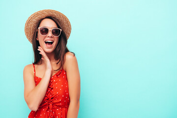 Young beautiful smiling female in trendy summer red dress. Sexy carefree woman posing near blue wall in studio. Positive brunette model having fun in hat and sunglasses. Screams and shouts