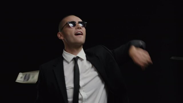 Young happy caucasian businessman in business suit and sunglasses dancing among falling dollar bills in slow motion isolated on black background. Hit a big jackpot. Successful cryptocurrency investor