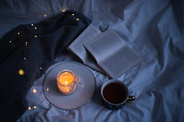 Burning scented candle on ceramic tray with open paper book and knit sweater cloth in bed. Winter cozy hygge atmosphere with cup of black coffee at home. Coziness.