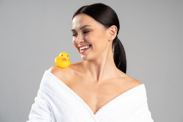 Obraz na płótnie Canvas Cute girl with dark hair and light nude make-up wearing white bath robe, looking at the rubber duck and smiling over the white studio background. Beauty photo concept