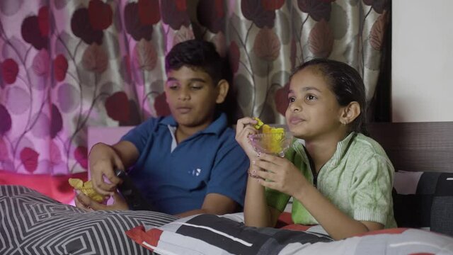 Kids at bedroom busy watching online web series on television or tv by having snacks during night - concept of unhealthy eating lifestyle, siblings bonding and relaxation
