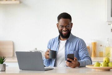 Smiling young black male with beard in glasses drinks coffee from cup, checks app on smartphone