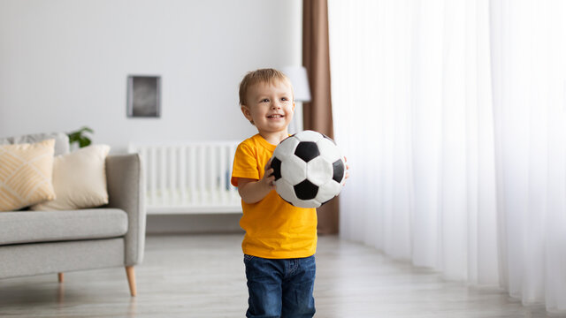 Favorite game. Happy toddler male kid holding football ball in hands and smiling while playing at home, panorama