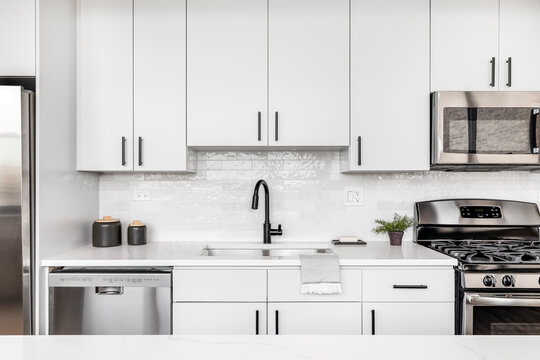 A beautiful white kitchen detail shot with a tiled backsplash, white cabinets, stainless steel appliances, and black hardware and faucet.