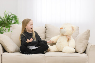 Photo of teddy bear and blonde girl looking on each other on the sofa.