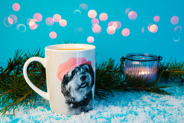 Scena con una tazza di caffè con l'immagine di un cagnolino sopra, circondata da rami di abete e con una candela sullo sfondo azzurro, e delle luci sfuocate. Bevanda. Inverno. Gennaio.  - obrazy, fototapety, plakaty