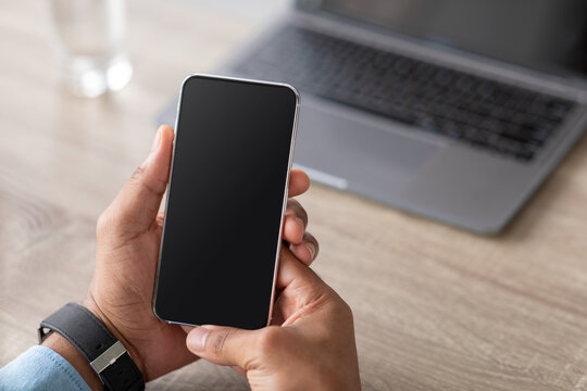 Unrecognizable millennial african american businessman holding smartphone with blank screen on workplace - Powered by Adobe