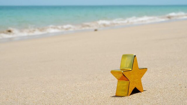 Little golden five-pointed star toy stand on sand of tropical island, selective focus shot. Small waves washing beach on background. Beautiful green-blue colours of crystal clear sea