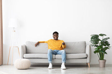 Joyful african american guy sitting on couch, turning on TV