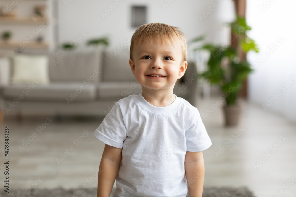 Wall mural portrait of adorable toddler boy wearing white t-shirt and smiling, posing to camera in living room 