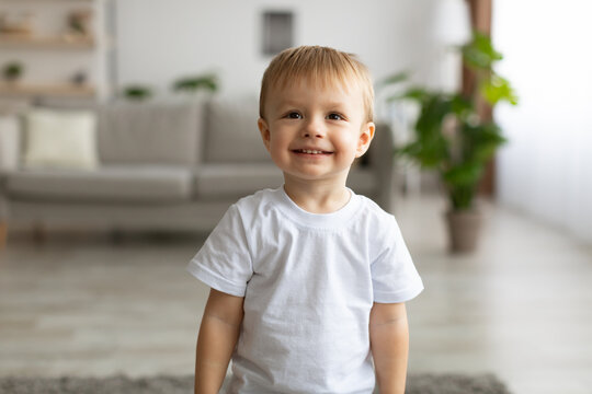 Portrait of adorable toddler boy wearing white t-shirt and smiling, posing to camera in living room interior