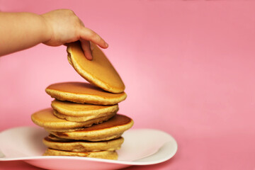 A child takes a pancake from a stack, pink background. American delicious pancakes. Close up, copy space