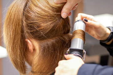 Fototapeta premium Hands of female hairstylist curls hair client with a curling iron in a hairdressing salon, close up