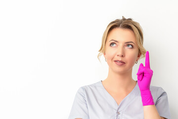 Naklejka premium Portrait of young caucasian doctor woman wearing rubber gloves in medical uniform with one finger up, looking up against a white background, copy space