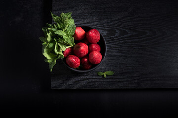 Fresas enteras con hojas de menta en un recipiente negro sobre una mesa de madera negra y fondo...