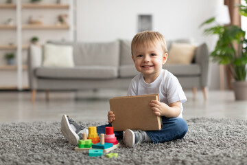 Adorable toddler boy holding book while playing with stacking and sorting toy, sitting on carpet and smiling at camera