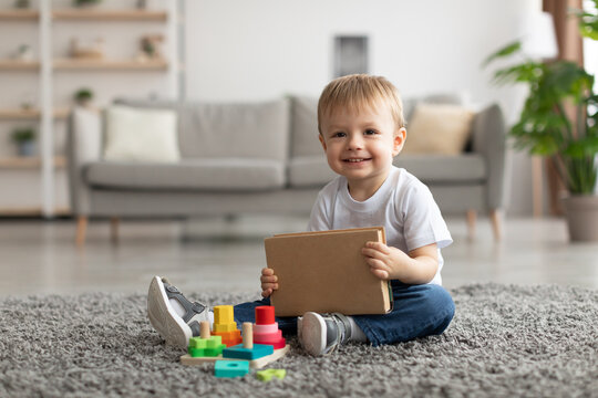 Adorable Toddler Boy Holding Book While Playing With Stacking And Sorting Toy, Sitting On Carpet And Smiling At Camera