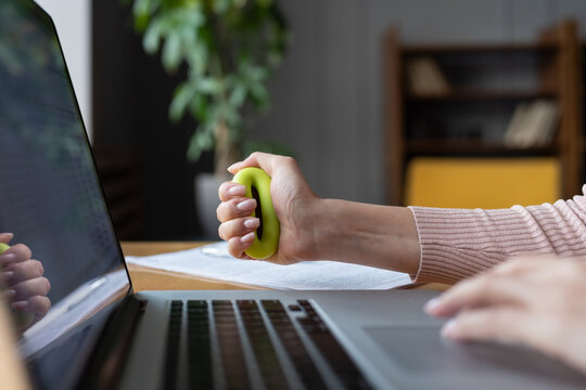 Closeup of woman office worker gripping rubber expander in hand while typing on laptop. Female doing exercises for wrist diseases prevention while working on computer. Tunnel syndrome injury concept
