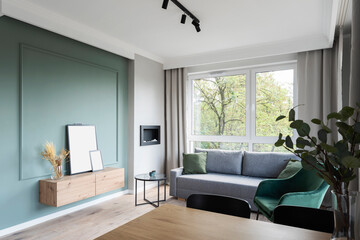 Interior of living room with stylish green wall, big window,comfortable sofa and frame with mock up. New apartment with modern furniture in scandinavian style.