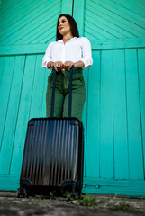 Young woman wearing green pants and a white shirt standing in front of a green door holding a black suitcase in both hands and waiting at a train station. Mention of business travel and vacations.