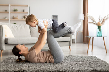 Fototapeta na wymiar Sport activities with baby. Young mother exercising with her toddler son, balancing kid on knees, training on carpet