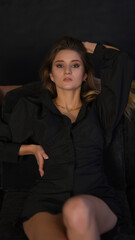 Portrait of a girl in a black dress. The girl is lying on the couch and looking at the camera. Thin figure. Black background.