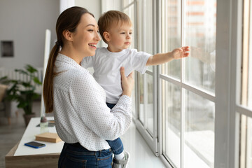 Happy mother holding toddler son on hands, standing by window showing him world outside, kid boy...